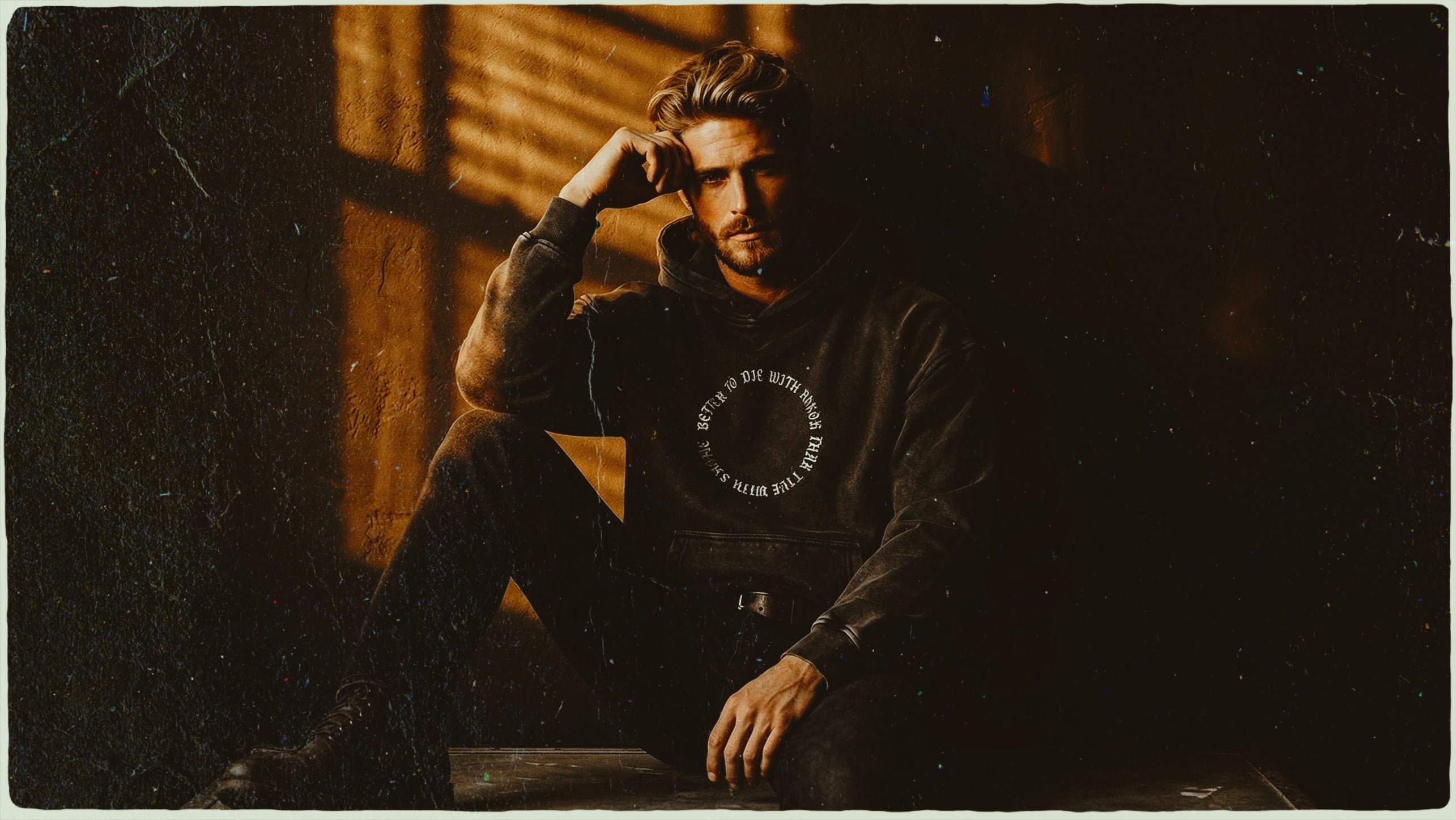 Man in a dark hoodie with circular text, sitting in dramatic shadow and light.