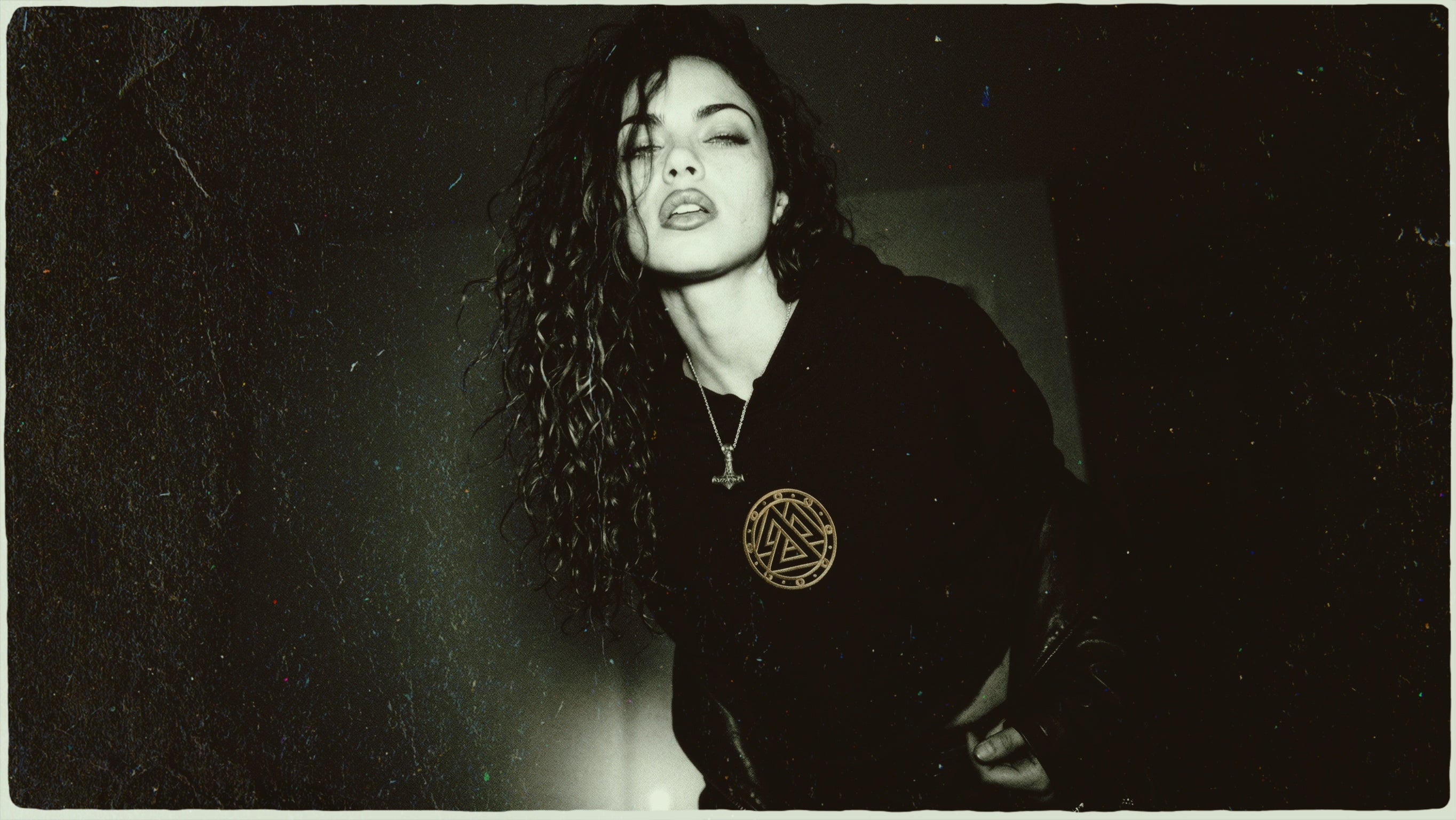 Woman with curly hair wearing a black hoodie with a gold Valknut symbol and a silver Mjolnir necklace.