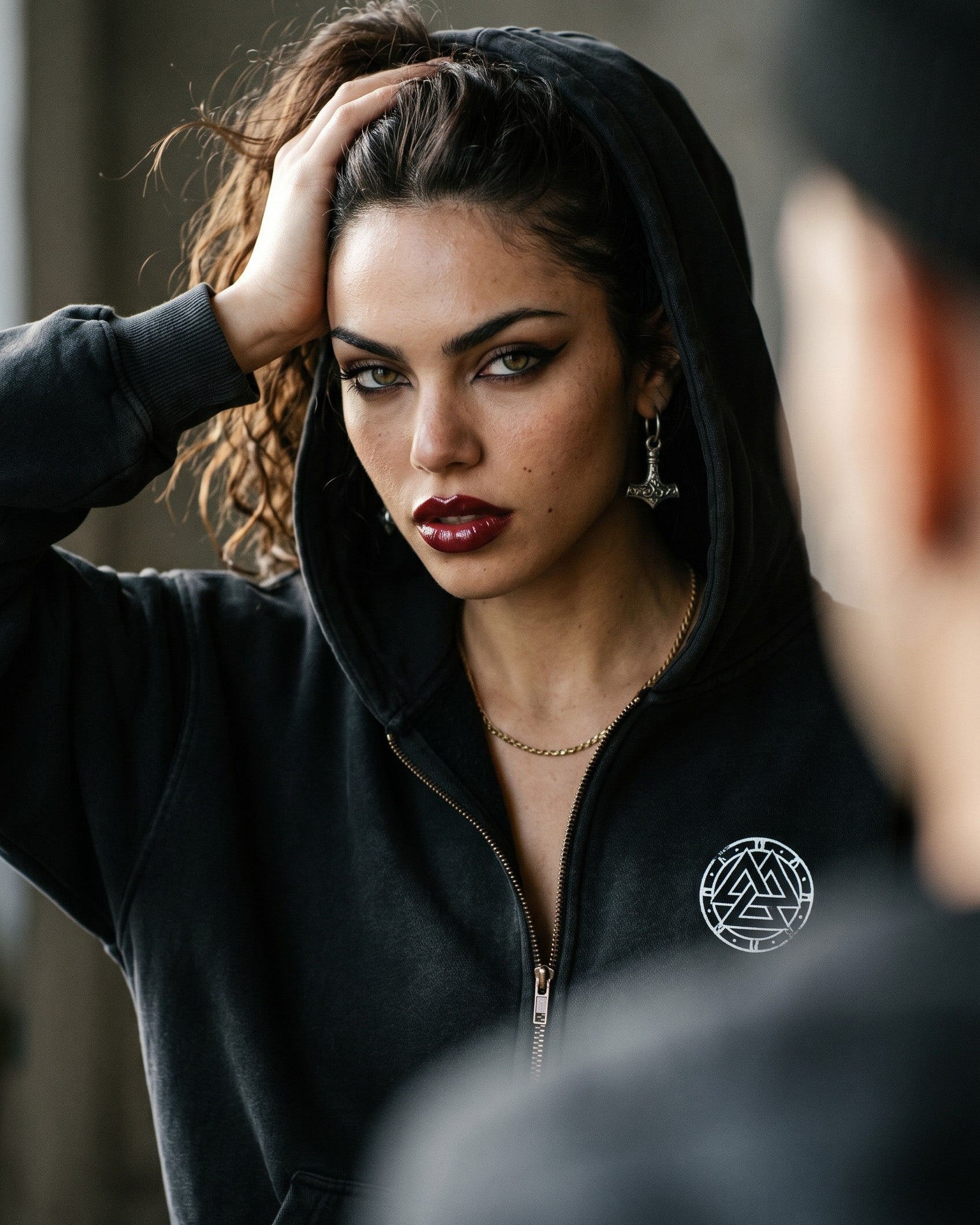 Woman in a sun-faded black Ragnarok hoodie with a Valknut logo, Mjolnir earrings, and dark red lipstick.